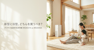 木の温もりを感じる明るいリビングで、母親と子どもがラグに座って本を読み、窓際には白いスリム型の加湿空気清浄機が設置されている。画像左側には「新型と旧型、どちらを買うべき？」と「ダイキン加湿空気清浄機 MCK557A vs MCK556A」の文字があり、2機種の購入を迷う人に向けた比較テーマを表現している。